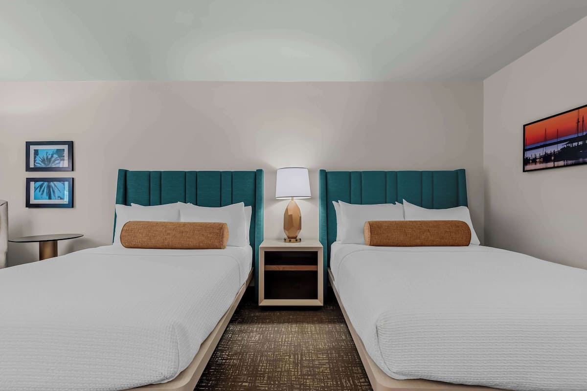 A hotel room with two beds, a nightstand with a lamp, a small table, and framed pictures on the walls.
