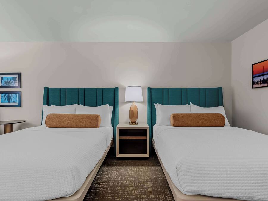 A hotel room with two beds, a nightstand with a lamp, a small table, and framed pictures on the walls.