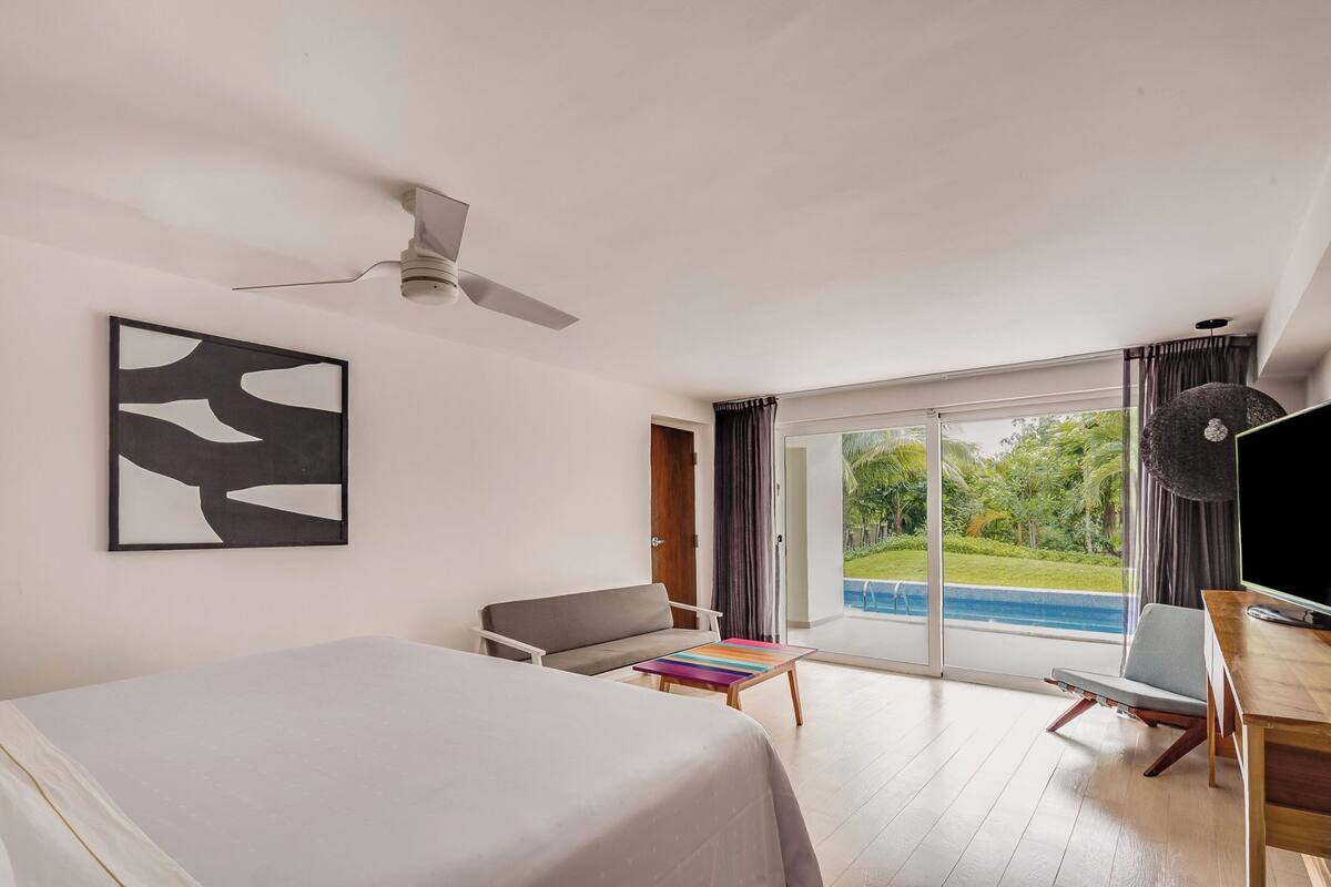 A modern hotel room with a bed, a desk, a chair, a ceiling fan, a large window with a view of a swimming pool, and a painting on the wall.