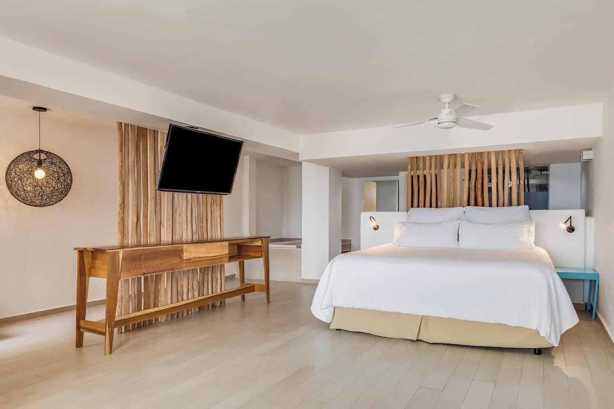 A modern bedroom with a large bed, a wooden bench, a flat-screen TV, and a ceiling fan.