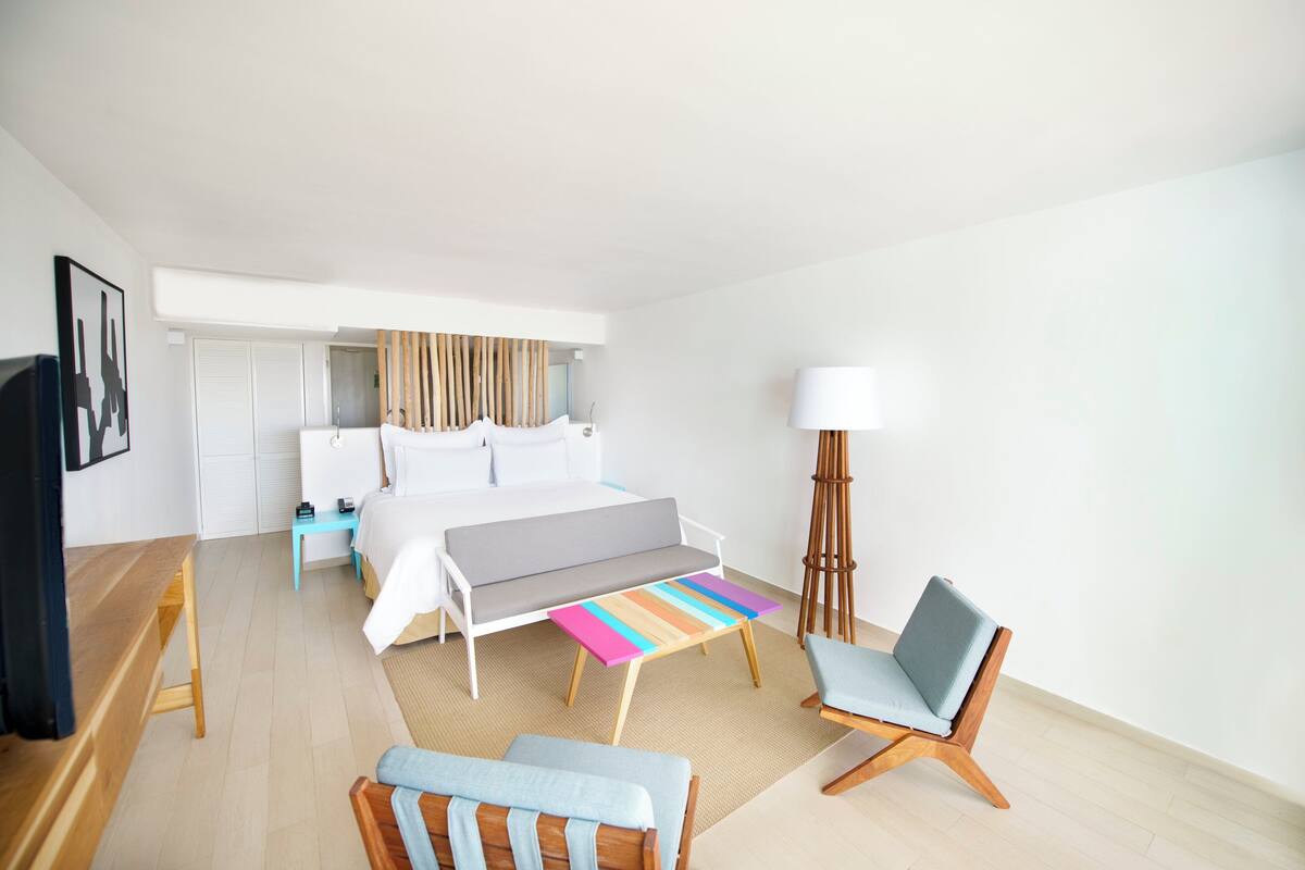 A modern hotel room with a bed, a desk, a chair, a bedside table, and a lamp.