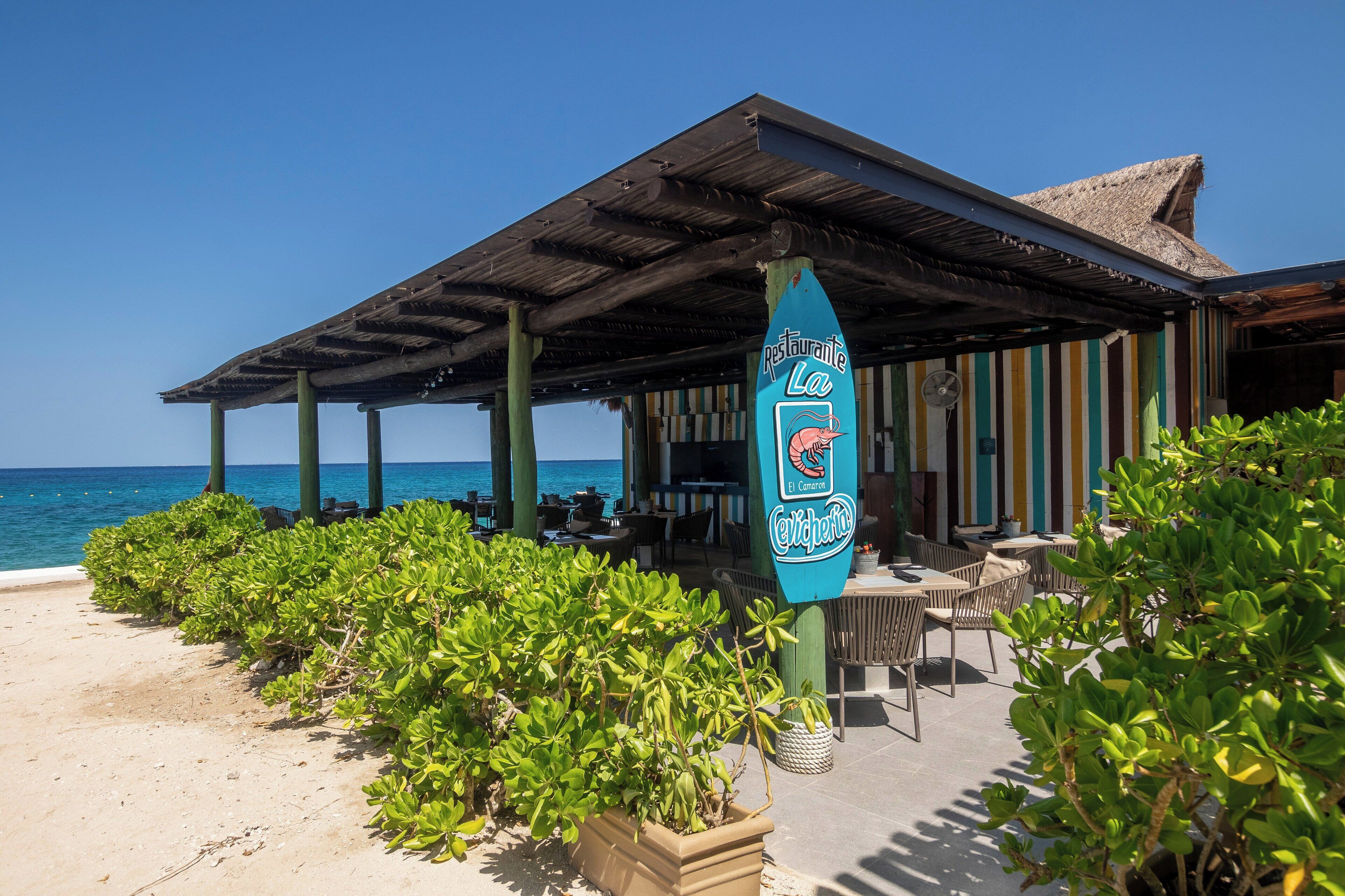 A beachside restaurant with outdoor seating, a thatched roof, and a view of the sea.