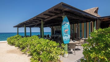 A beachside restaurant with outdoor seating, a thatched roof, and a view of the sea.