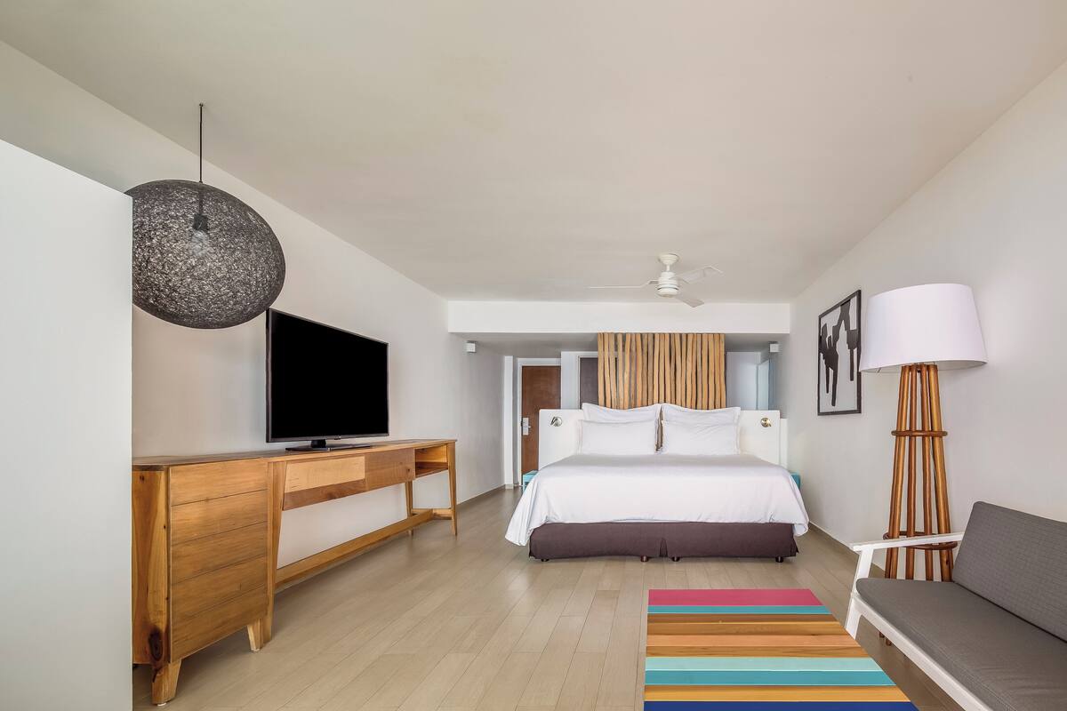 A modern hotel room with a wooden desk, a flat-screen TV, a bed with white bedding, a colorful striped rug, and a standing lamp.