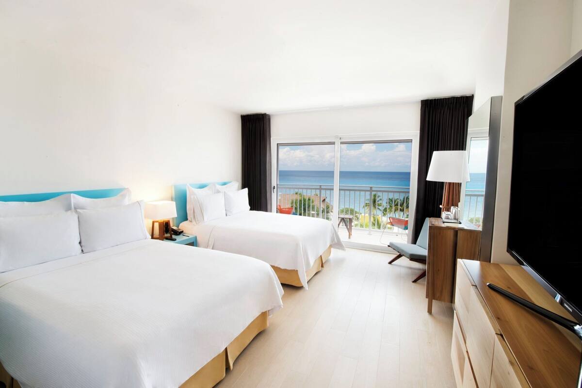 A hotel room with two beds, a TV, a desk, and a balcony with a view of the ocean.