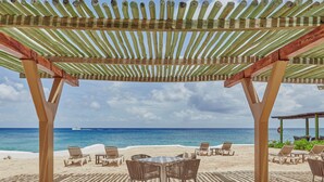 A beachside patio with a wooden pergola, lounge chairs, and a table.