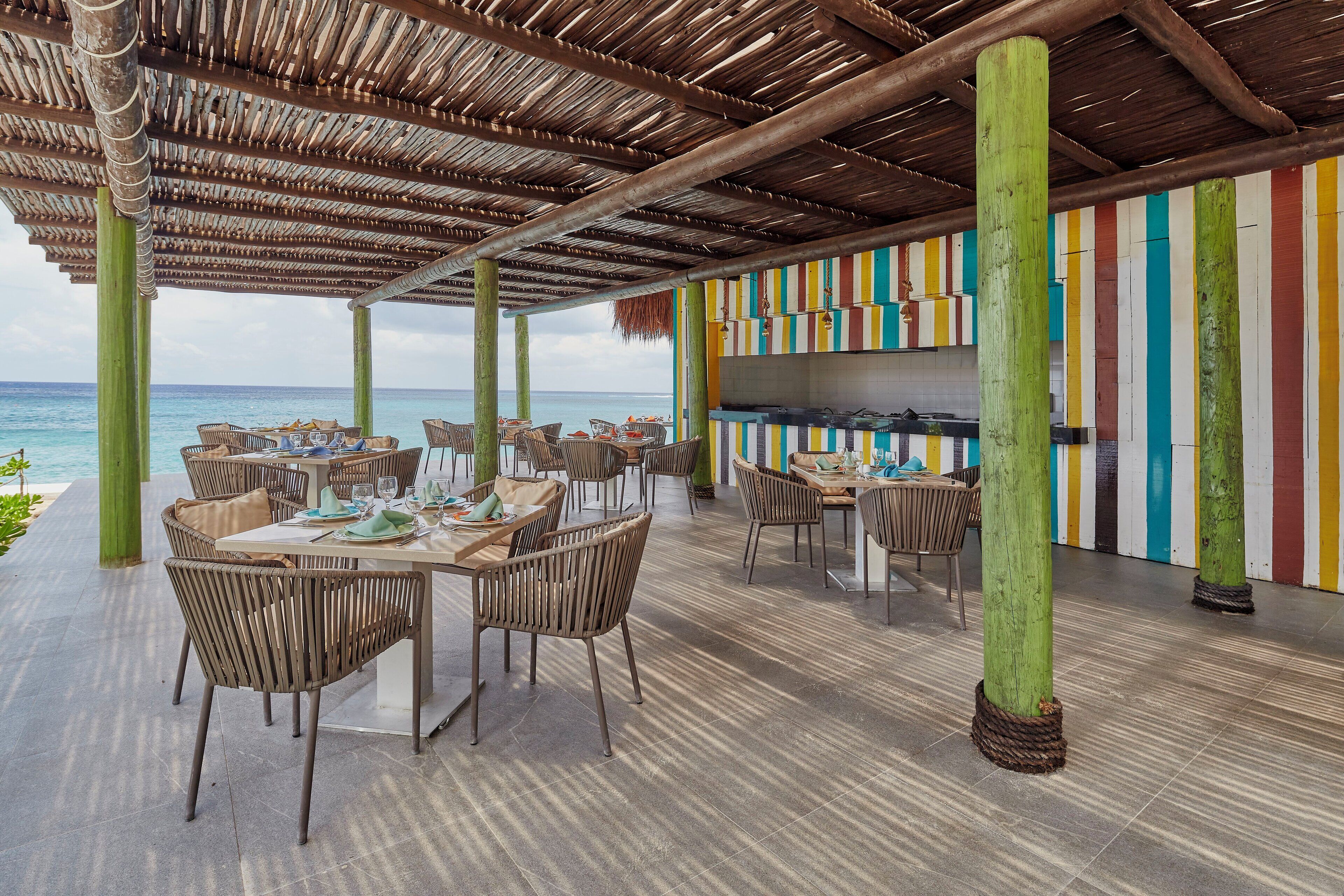 Un área de comedor al aire libre con mesas y sillas, techo de paja y vista al océano.