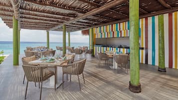 Un área de comedor al aire libre con mesas y sillas, techo de paja y vista al océano.