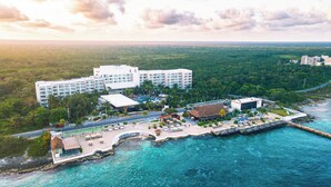An aerial view of a resort with a large hotel, a beach area with lounge chairs, and a pier extending into the water.