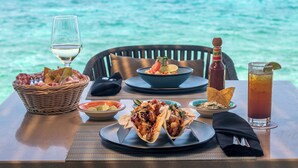 A dining table with a basket of bread, a bowl of salsa, two plates of tacos, a glass of white wine, a bottle of hot sauce, a glass of iced tea, and a napkin.