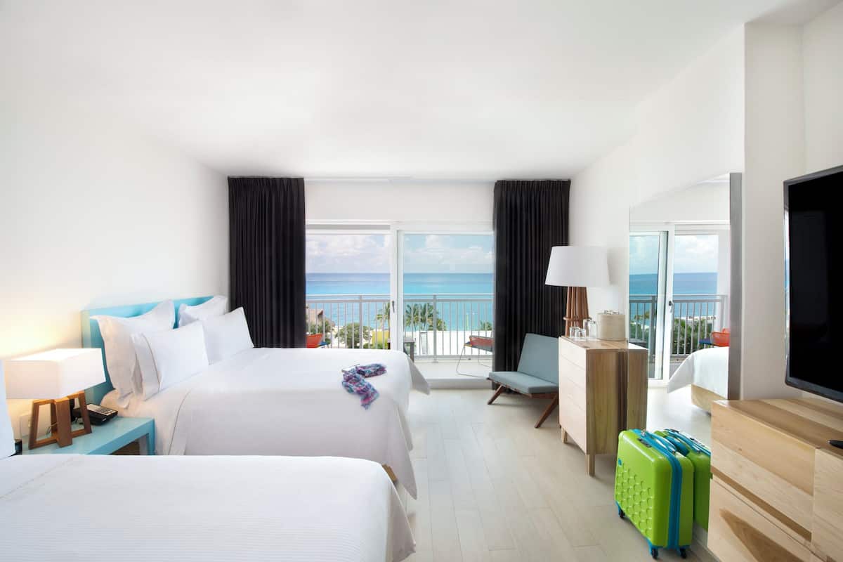 A hotel room with two beds, a TV, a desk, a chair, and a view of the ocean.