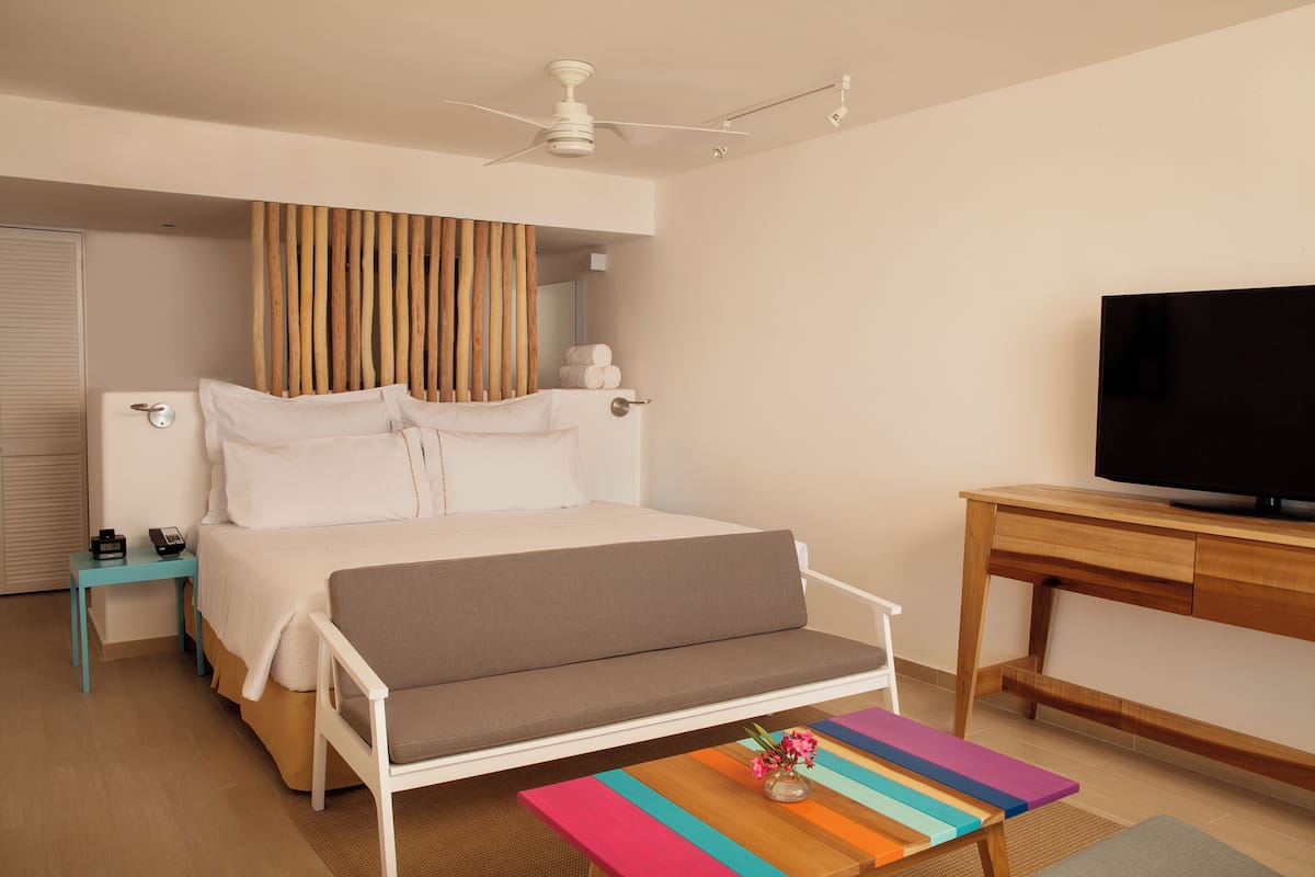 A modern hotel room with a bed, a TV, a wooden desk, and a colorful striped table.