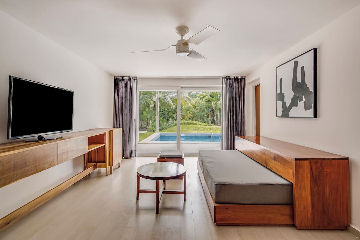 A modern living room with a flat-screen TV, wooden furniture, a ceiling fan, and a view of a swimming pool.