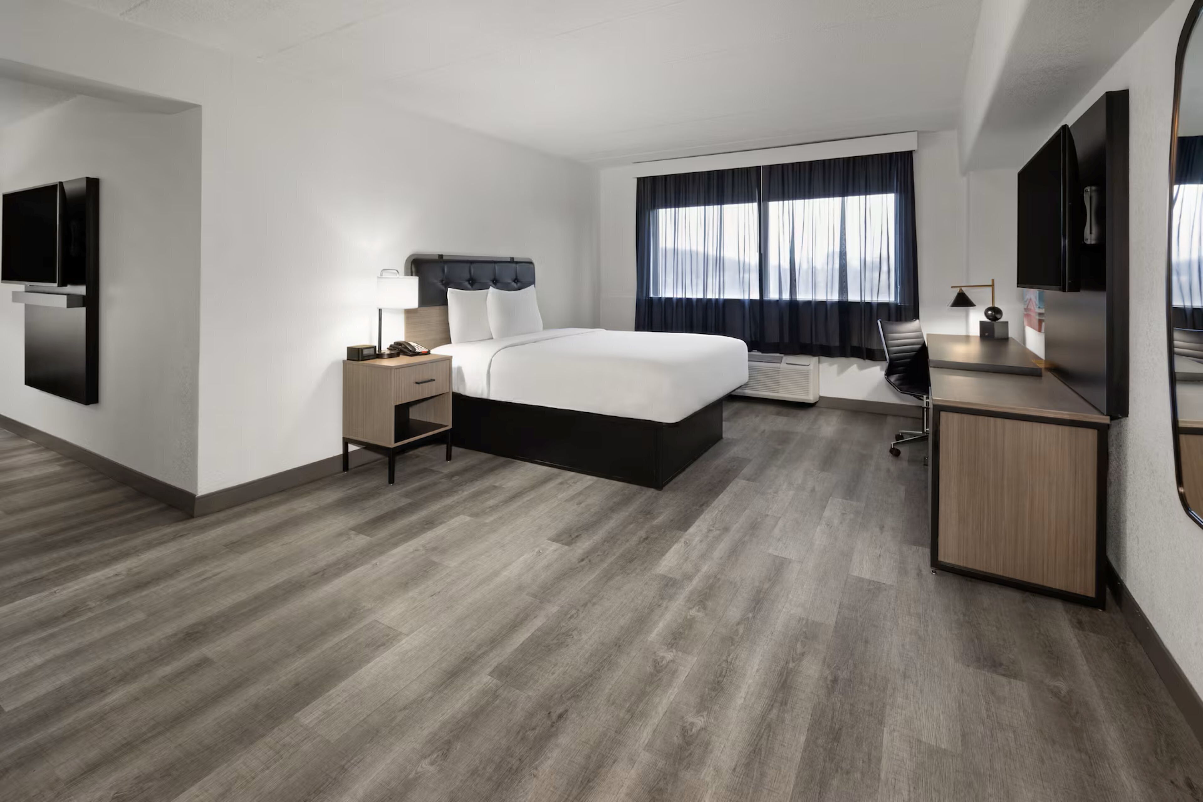 A modern hotel room with a bed, desk, chair, TV, and a large window with curtains.