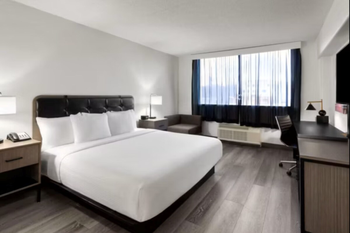 A modern hotel room with a large bed, a desk, a chair, and a window with curtains.