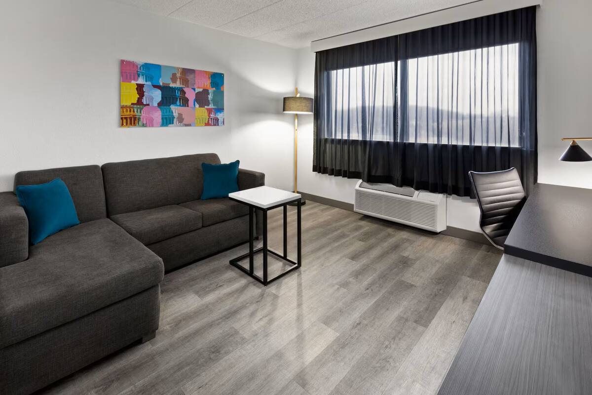 A modern living room with a grey sofa, a small white table, a black chair, a large window with black curtains, and a colorful abstract painting on the wall.