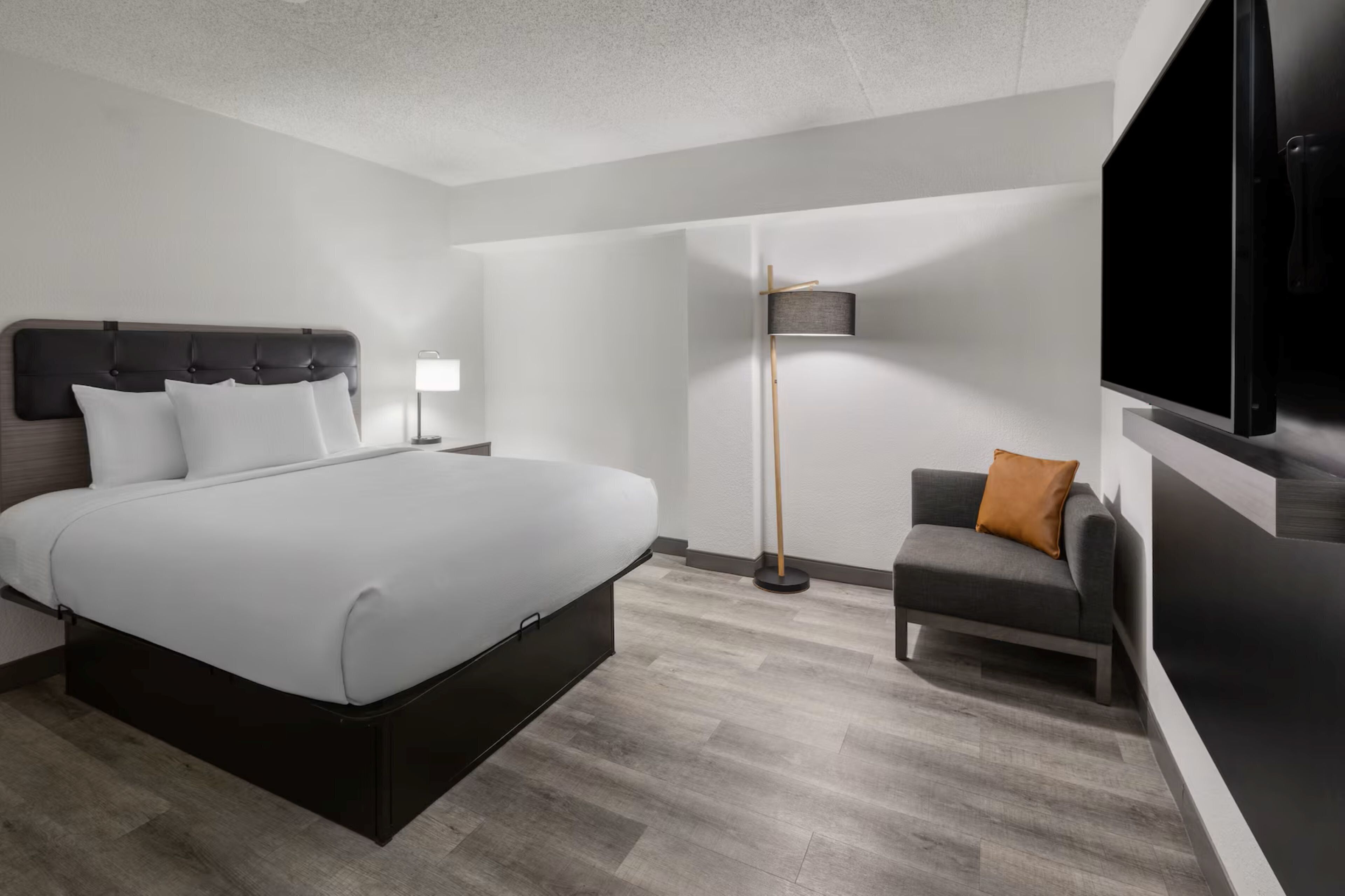 A modern hotel room with a large bed, a desk, a chair, a TV, and a floor lamp.