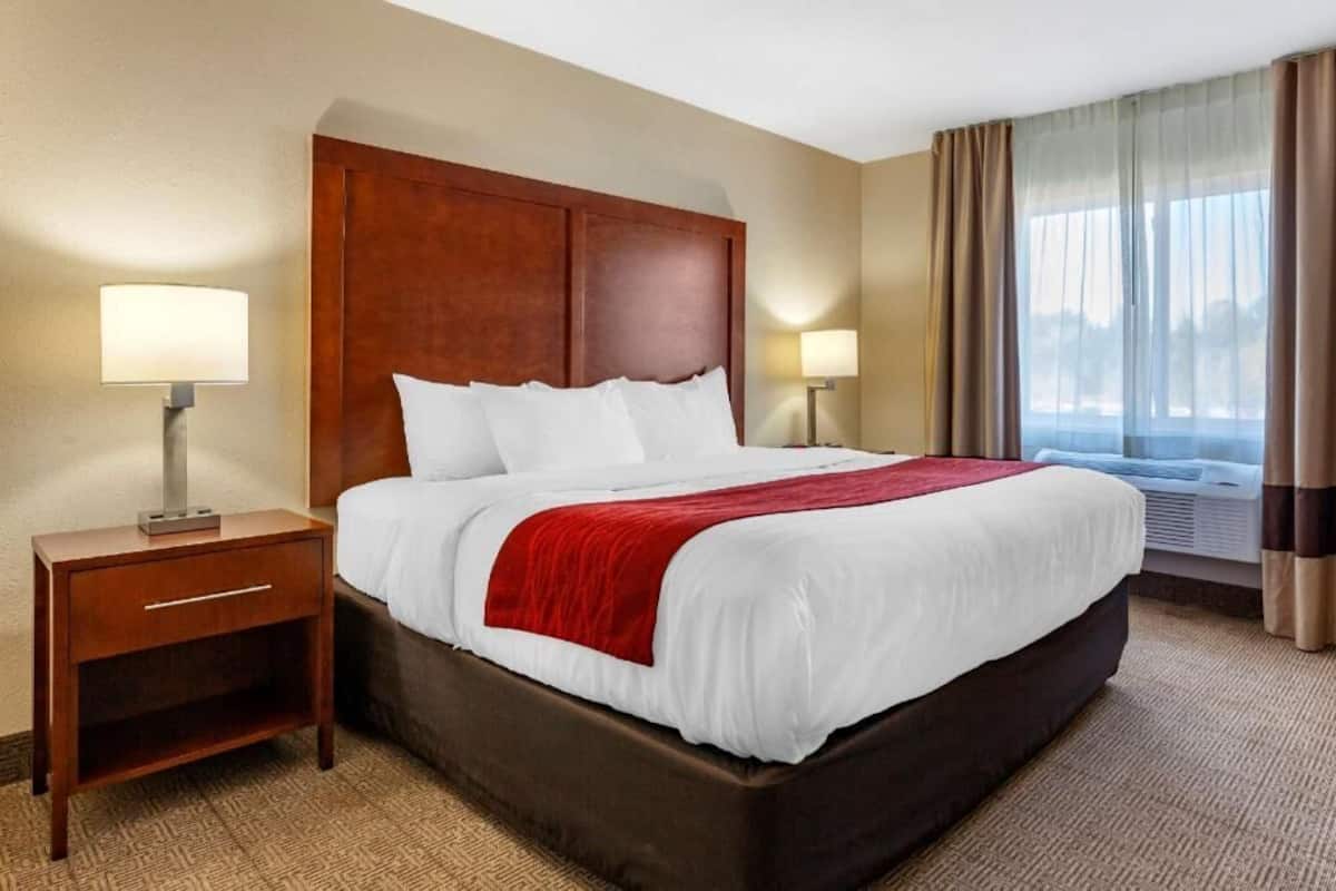 A hotel room with a large bed, two bedside tables, a nightstand, a window with curtains, and an air conditioning unit.