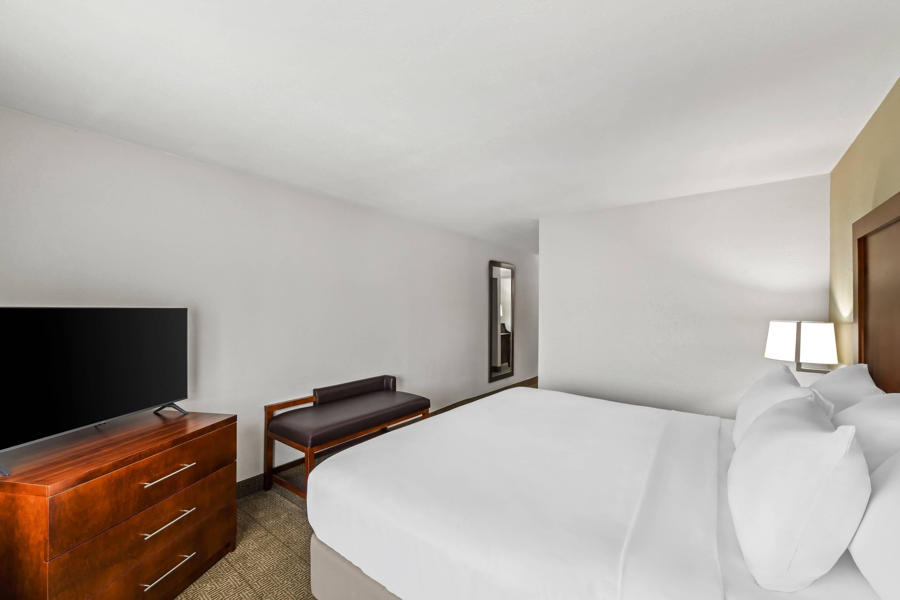 Una habitació d'hotel amb un llit, una televisió sobre una cómoda de fusta, una cadira, una tauleta de nit i una làmpada.