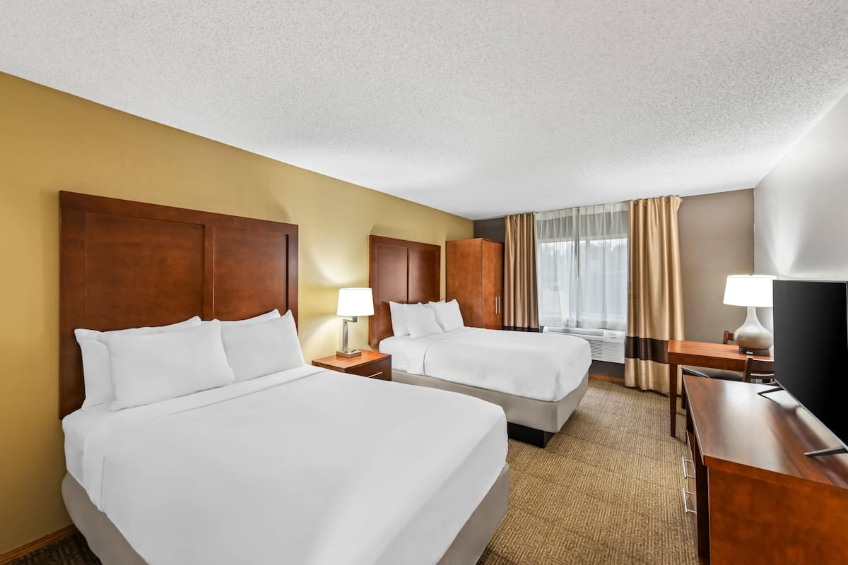 A hotel room with two beds, wooden headboards, a desk, a TV, and a window with curtains.