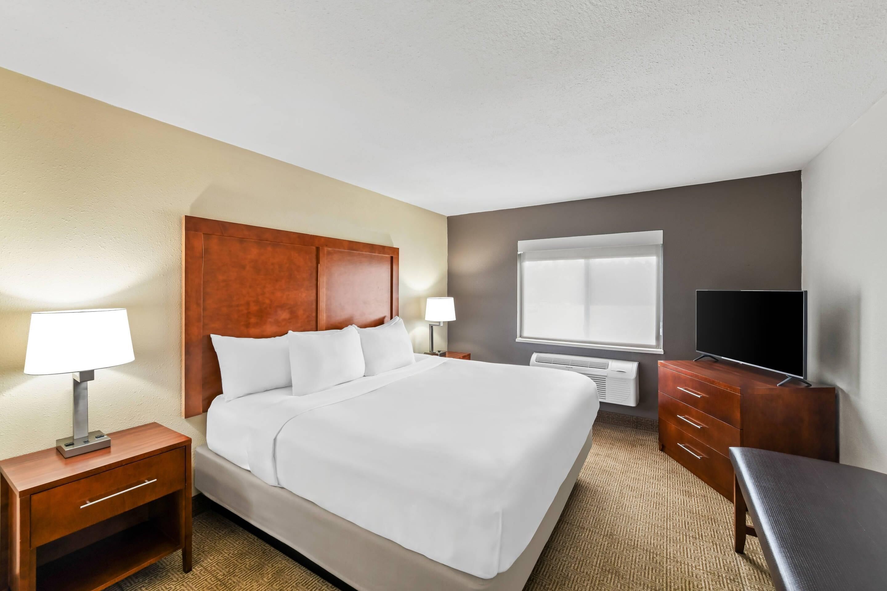 Habitación de hotel con una cama grande, cabecera de madera, dos mesitas de noche con lámparas, un televisor de pantalla plana y una ventana con persianas.