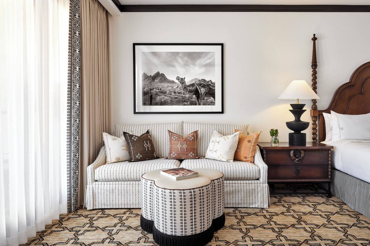 Een hotelkamer met een gestreept sofa, een rond poef, een ingelijst kunstwerk aan de muur, een nachtkastje met een lamp en een kamerkleed met een patroon.