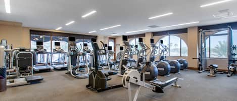 A gym with various cardio and strength training equipment, large windows, and a carpeted floor.
