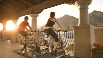 Dos personas hacen ejercicio en bicicletas estáticas bajo una estructura cubierta, con vistas a montañas al fondo.