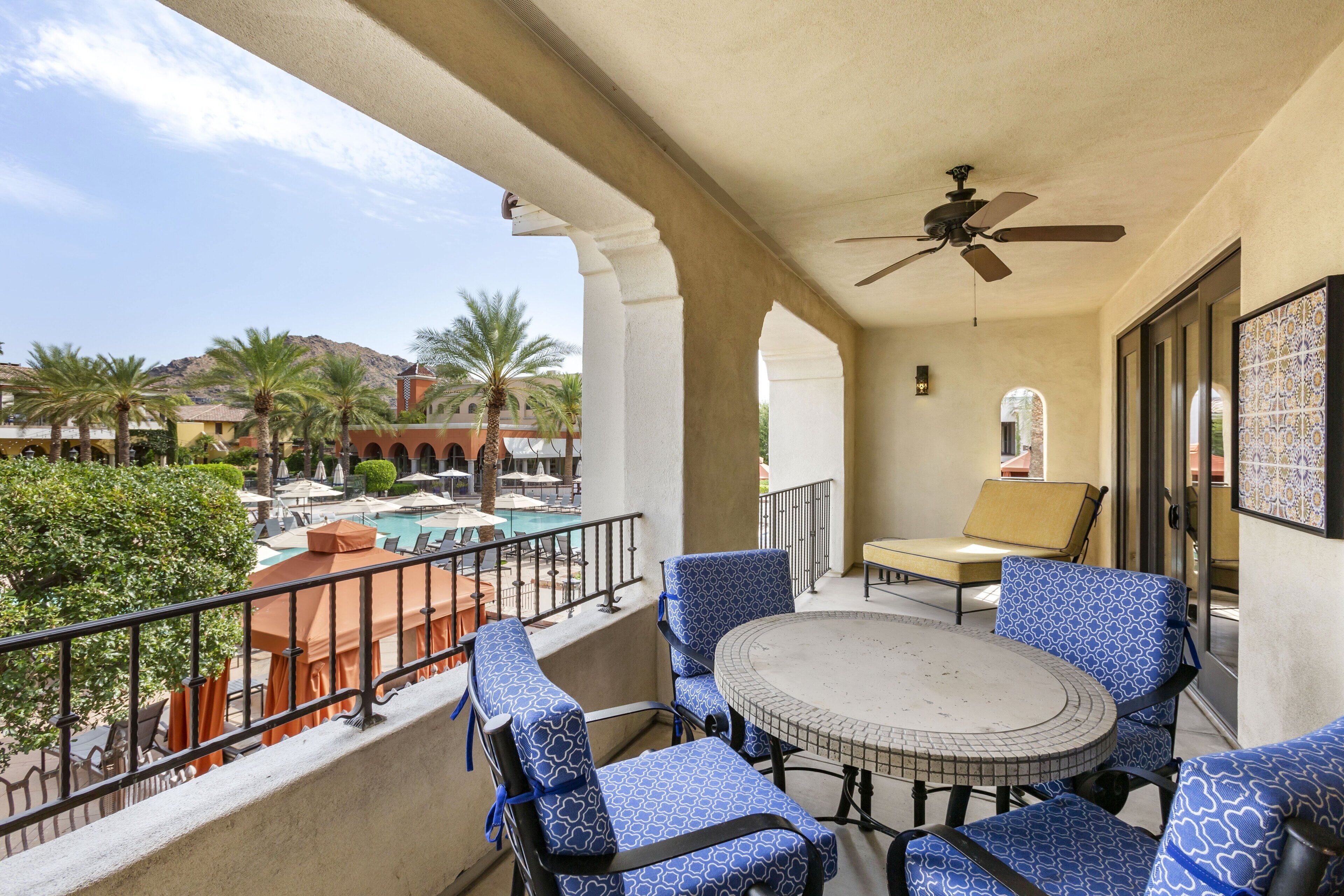 A balcony with a round table and blue patterned chairs, overlooking a pool and palm trees.