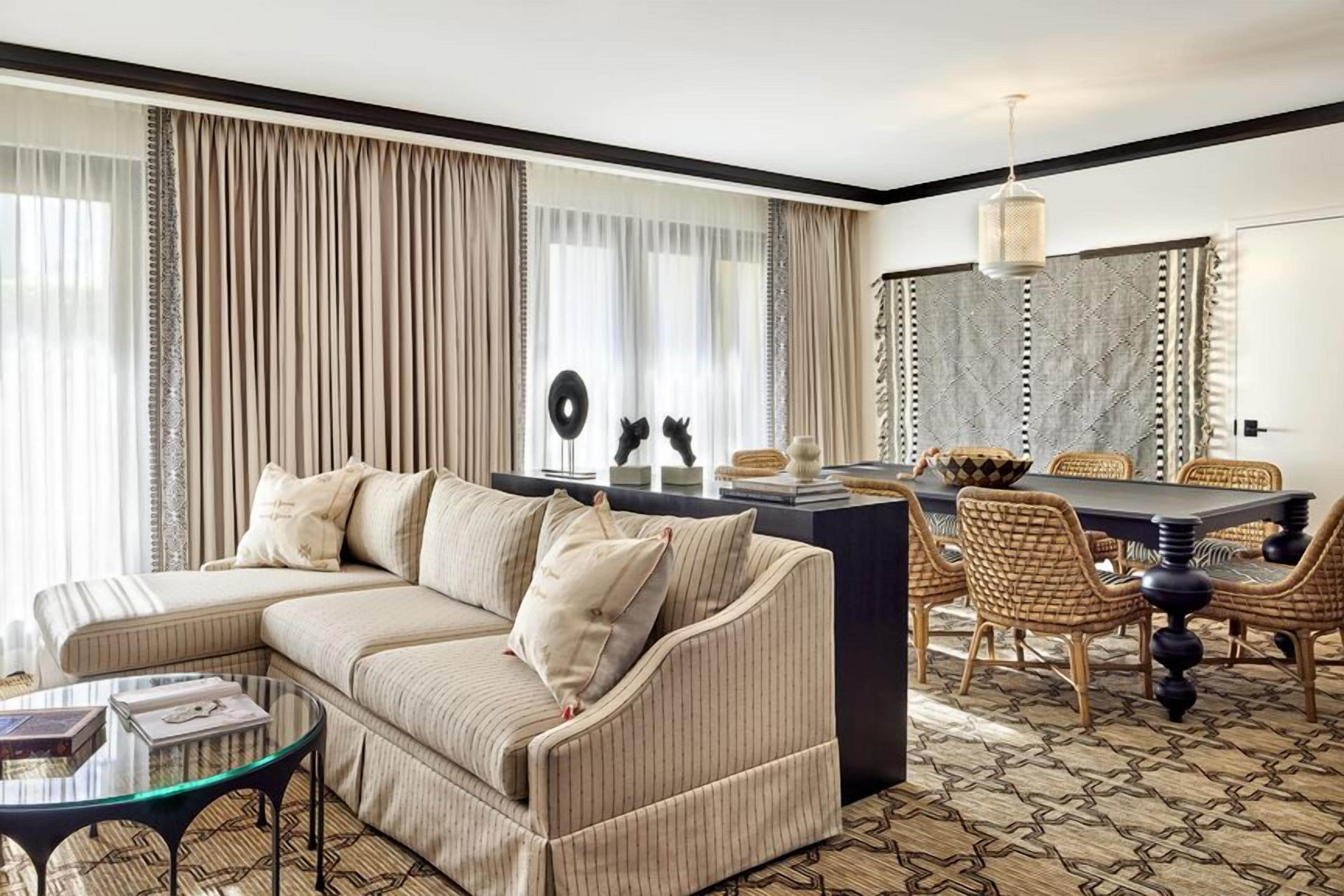 Ein Wohnzimmer mit einer Couch, Couchtisch, Esstisch und Stühlen. Es gibt ein großes Fenster mit Vorhängen, eine Chandelüster und einen gestreiften Boden.