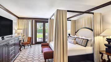 A hotel room with a four-poster bed, a television, a desk with a lamp, and a view of the outdoors.