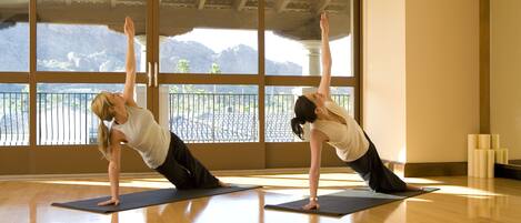 Dos personas realizando una postura de yoga sobre esterillas en una habitación con ventanas amplias y suelo de madera.