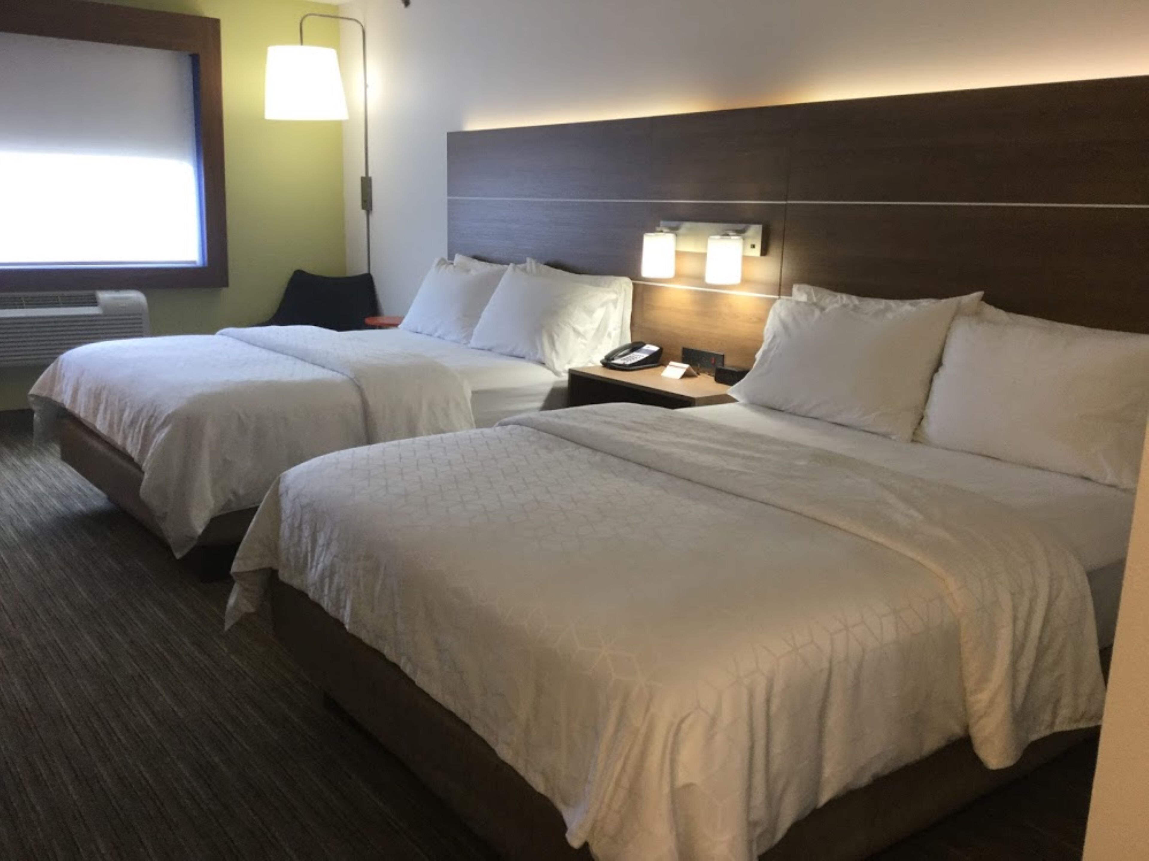 Habitación de hotel con dos camas, una mesita de noche con teléfono y lámpara, y una ventana con persianas.