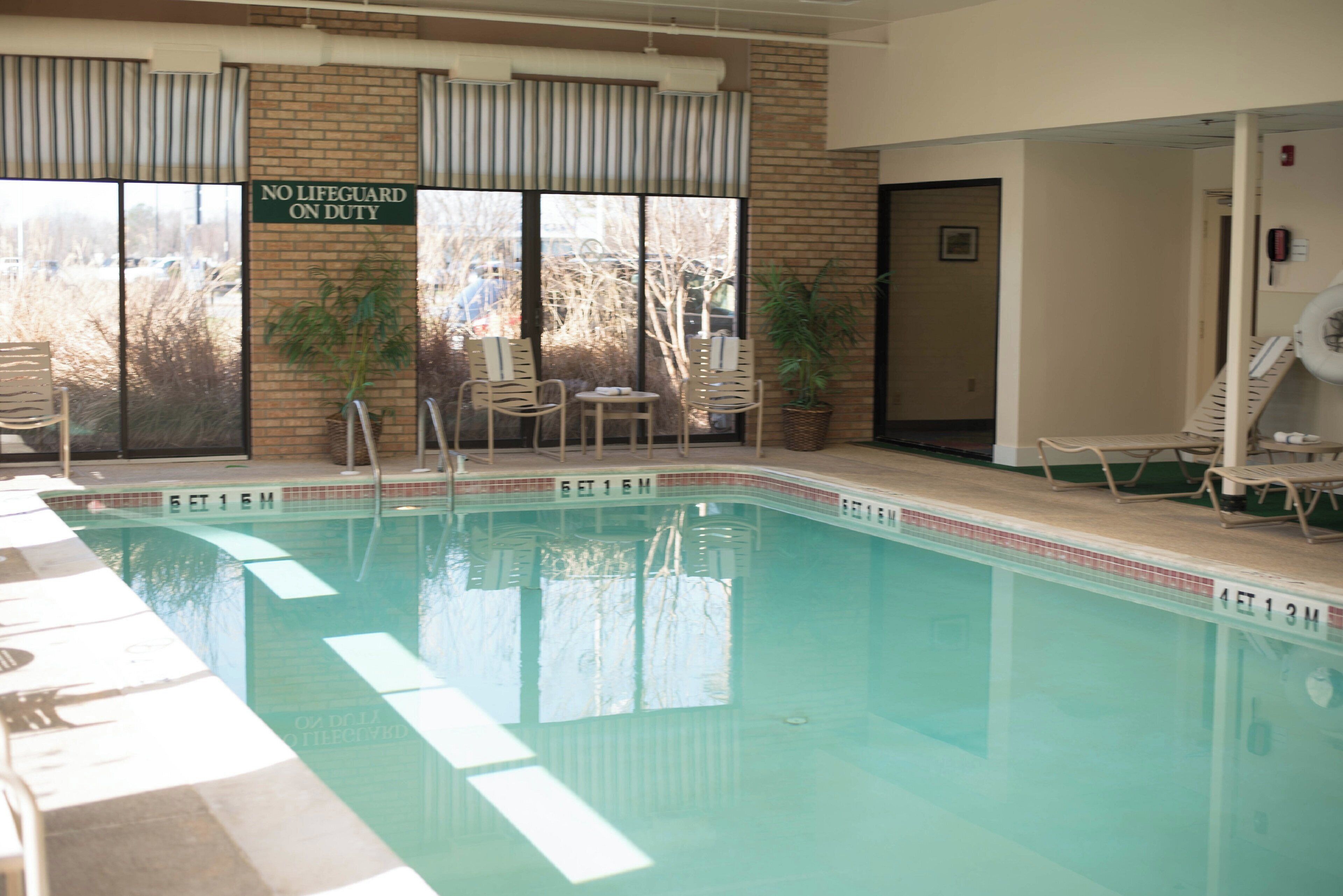 An indoor swimming pool with a 'NO LIFEGUARD ON DUTY' sign, lounge chairs, and potted plants.