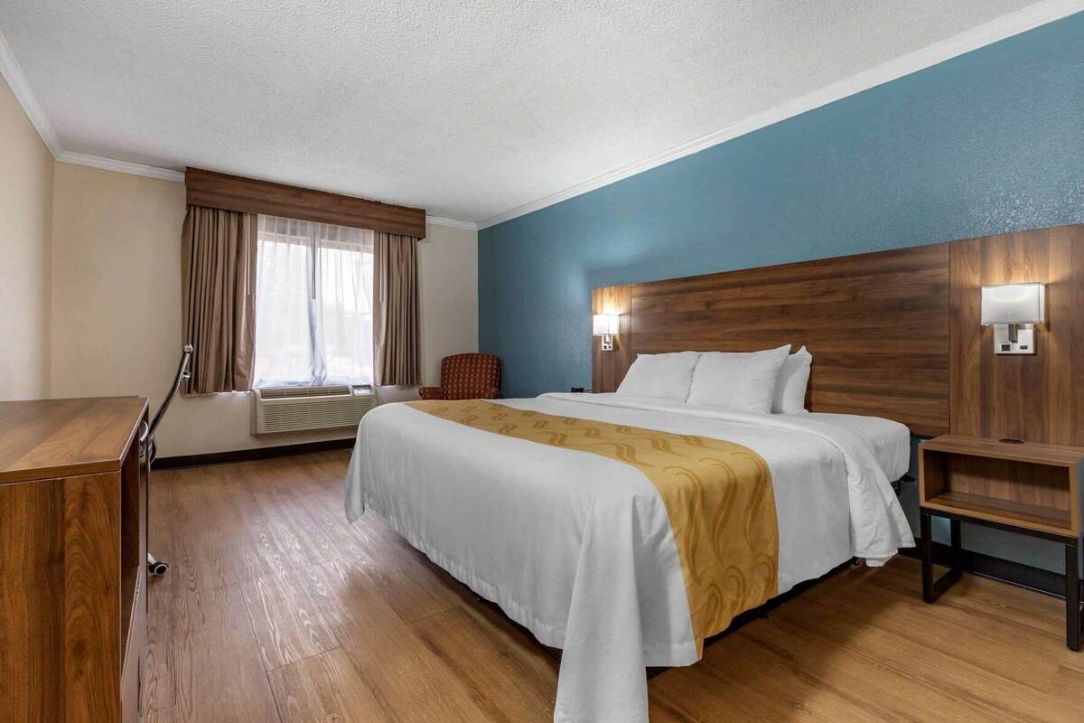 A hotel room with a large bed, a desk, a chair, a nightstand, and a wall-mounted lamp.