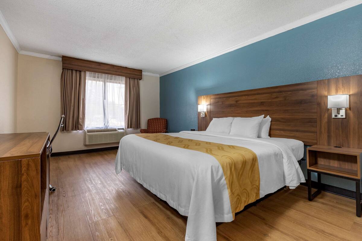 A hotel room with a large bed, a wooden desk, a chair, and a window with curtains.