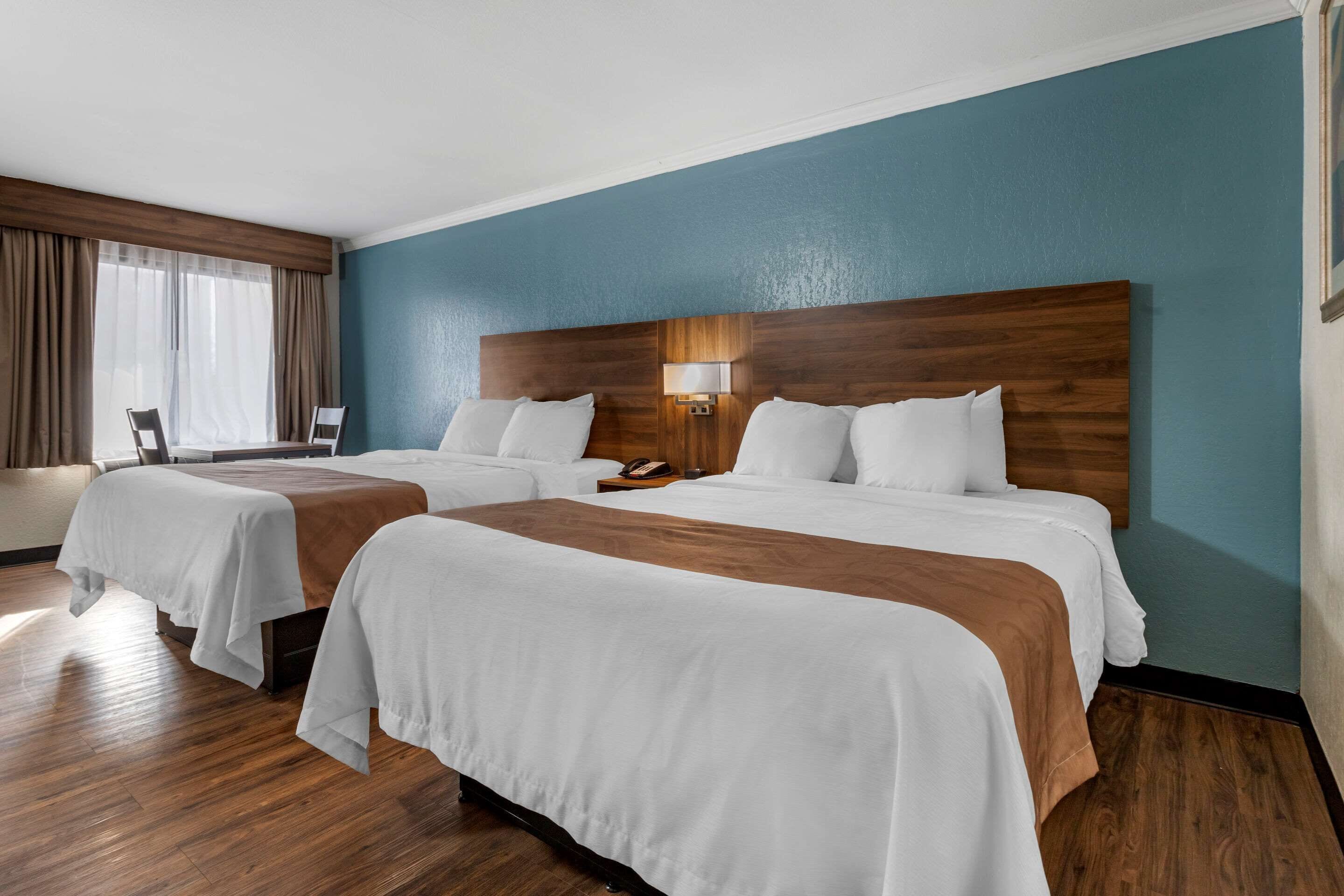 Une chambre d’hôtel avec deux lits, une tête de lit en bois, une table de chevet avec une lampe et une fenêtre avec des rideaux.