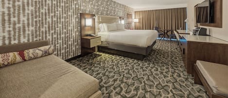 A hotel room with a patterned carpet, a bed, a desk, a chair, and a television.