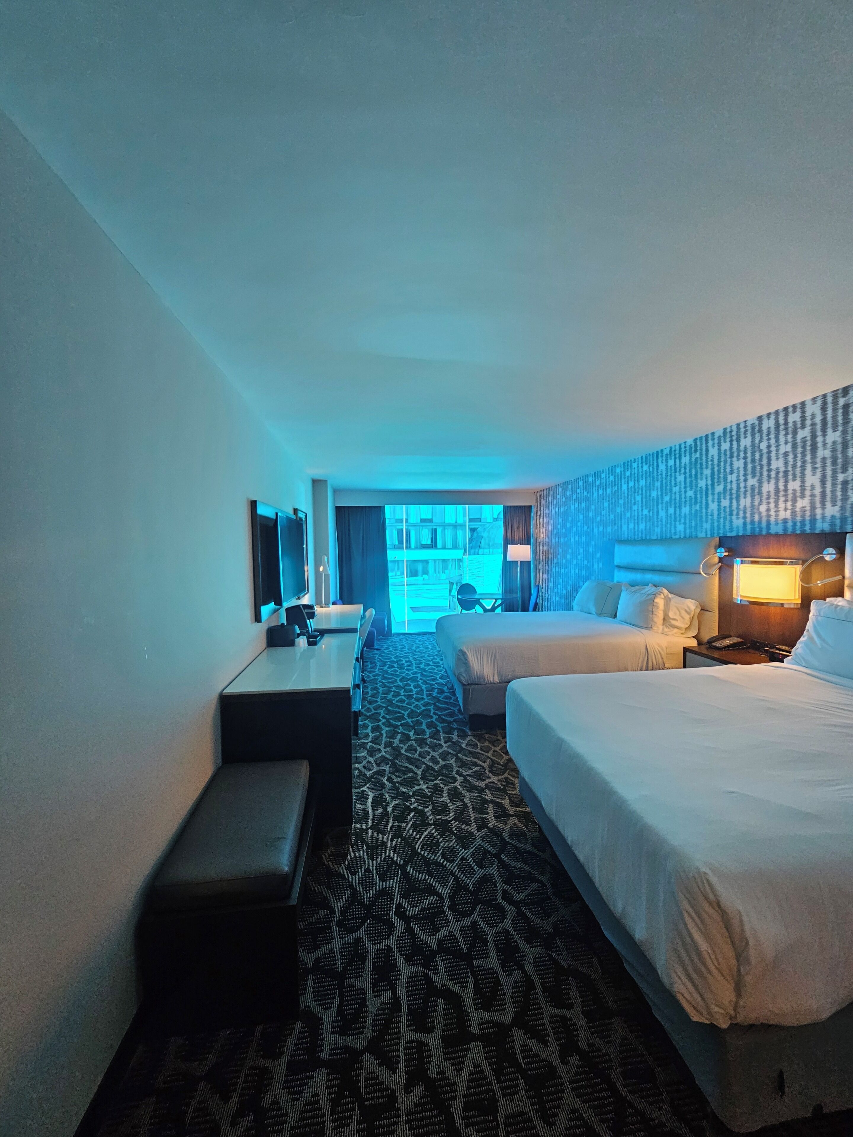 A hotel room with two beds, a desk, a chair, and a view of the outside.