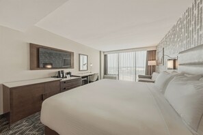 A hotel room with a large bed, a desk with a chair, a minibar, and a large window offering a view.