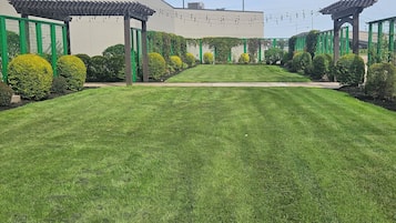Une pelouse bien entretenue, avec un chemin dégagé menant à l’entrée d’un bâtiment, le tout bordé d’arbustes soigneusement taillés et agrémenté d’une structure en forme de pergola.