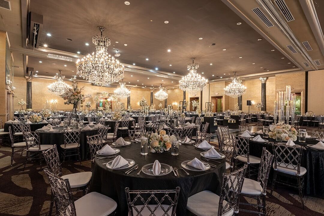 Salão de festas com mesas redondas preparadas para um evento formal, decorado com centros de mesa florais e elegantes louças.