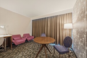A hotel room with a round wooden table, two pink armchairs, a blue patterned armchair, a desk with a lamp, and a patterned carpet.