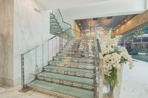 A grand staircase with ornate metal railings and glass steps, leading to a second-floor area with a chandelier and a large vase of white flowers.
