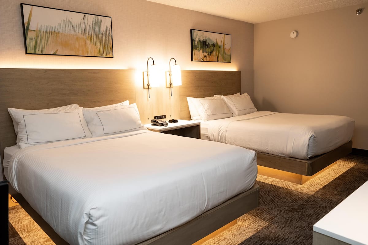 A hotel room with two beds, a nightstand with a phone and a remote control, and wall-mounted lamps.