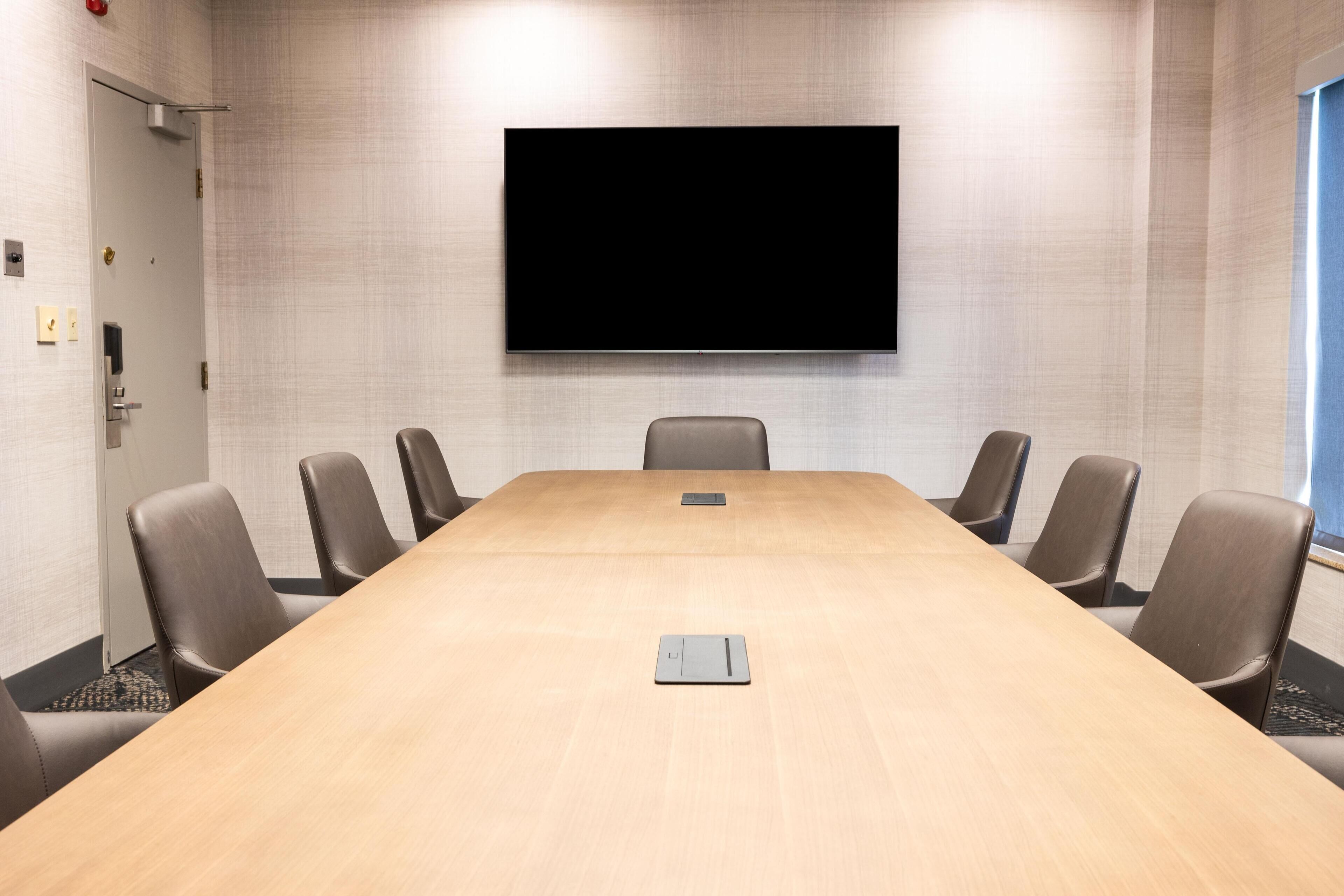 A conference room with a long table, six chairs, a flat-screen TV mounted on the wall, and a door on the left.