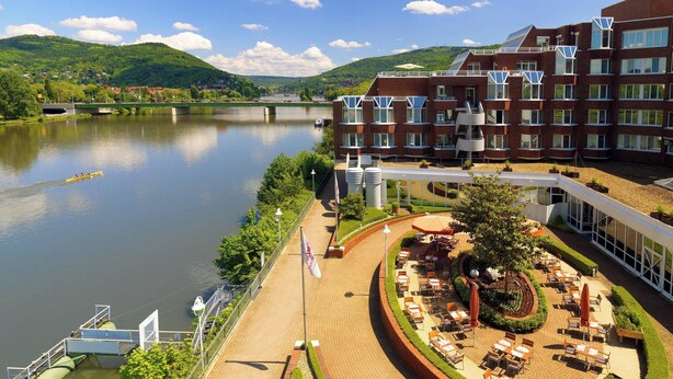 Un hotel con terraza junto al río, mesas al aire libre y vistas al río y las colinas.