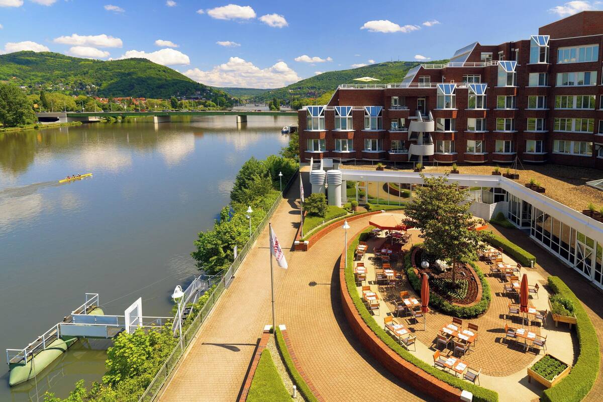 A hotel with a riverside terrace, outdoor seating, and a view of the river and hills.