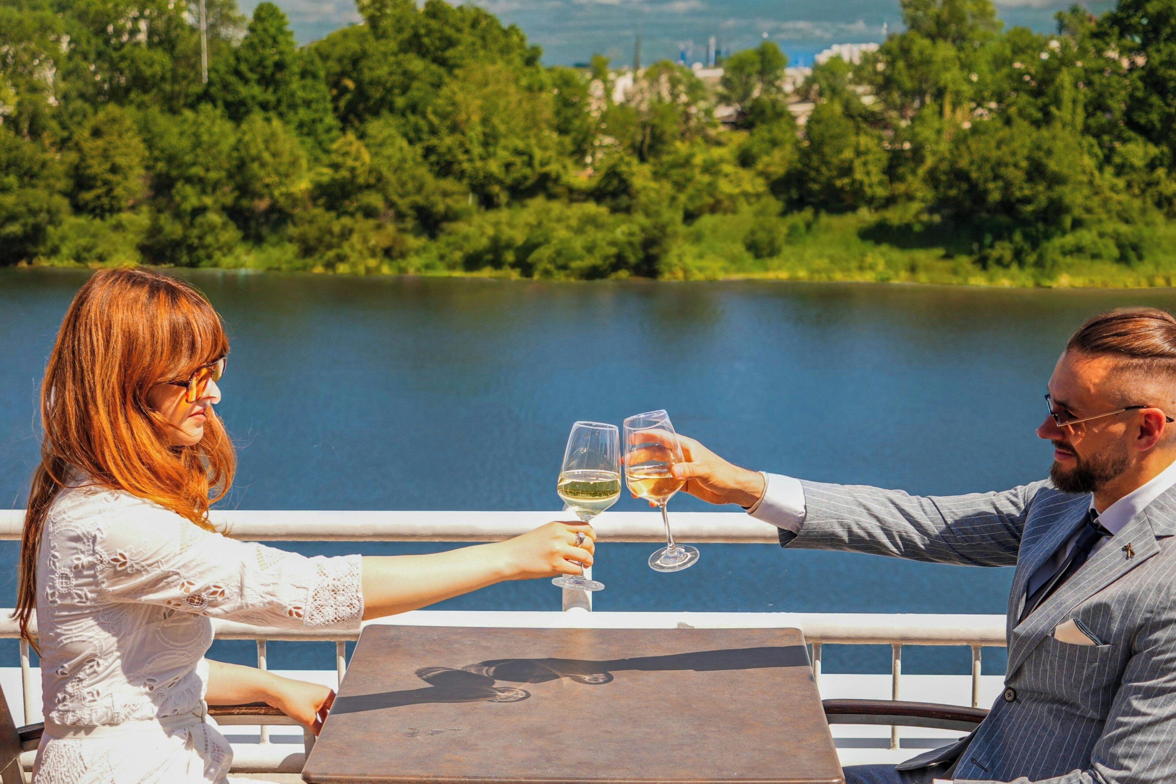 Um casal brindando com taças de vinho em um barco, com um lago e árvores ao fundo.