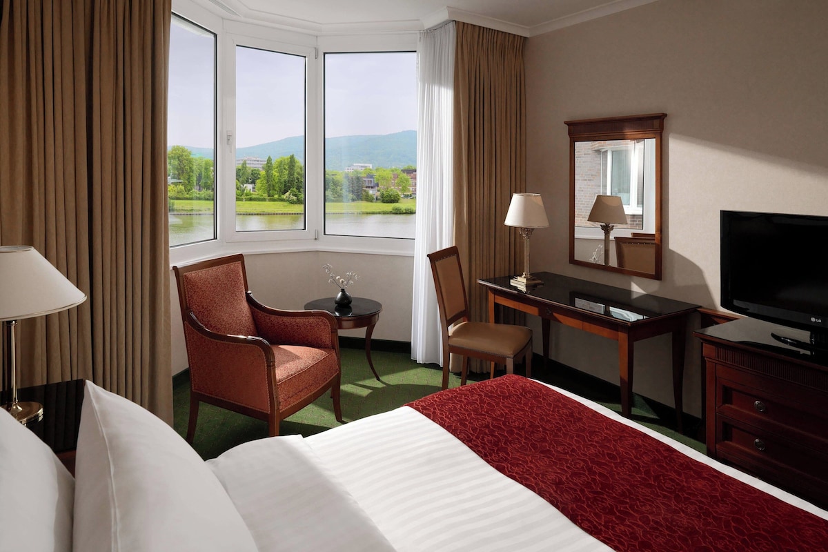 A hotel room with a bed, a desk, a chair, a TV, and a view of a lake and mountains.